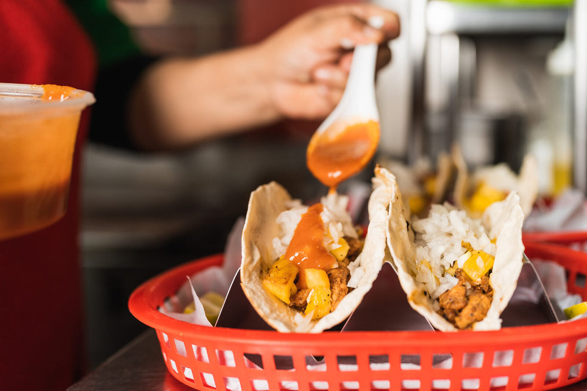 A cook pouring hot sauce over a basket of street tacos